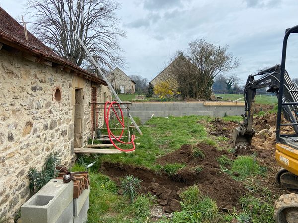 Mini-pelle creusant une tranchée de fondations près d’un mur en pierre