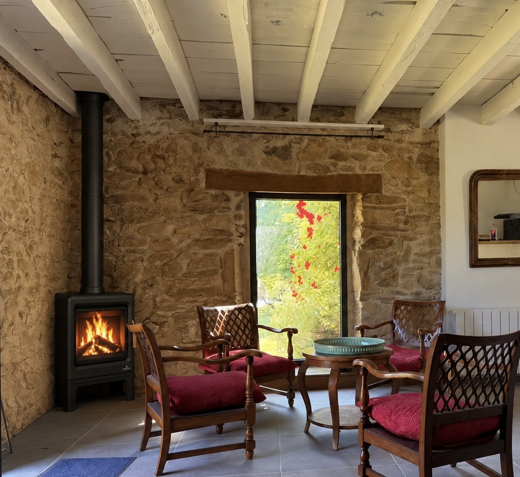 Le salon de La Bergerie : un poêle en fonte allumé contre le mur de pierre, quatre fauteuils à coussins rouges et une porte-fenêtre ouverte sur le jardin